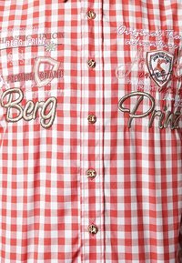 Camisa de cuadros rojos y blancos con texto y diseños bordados. Tiene botones brillantes de color dorado y un collar.