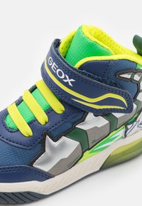 Mėlyni ir žali vaikų sportbačiai su geltonomis raištelėmis, Velcro dirželiu, pažymėtu "GEOX", ir žvaigždės formos sidabriniu bei žaliu šonu.