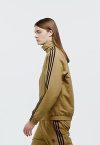 Khaki zip-up jacket with high collar, featuring black side stripes. Smooth fabric; model is shown walking against a white background.