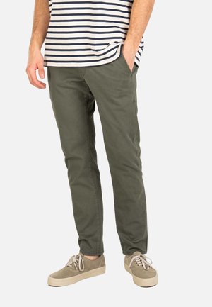 Hombre vistiendo pantalones ajustados color verde oliva, zapatillas beige y una camiseta blanca con rayas azules marinas, con una mano en el bolsillo.