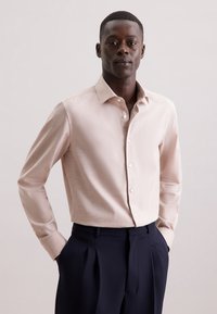 Camicia a maniche lunghe rosa chiaro con colletto button-down e patta frontale, abbinata a pantaloni blu scuro. Tessuto di texture morbida, vestibilità sartoriale.
