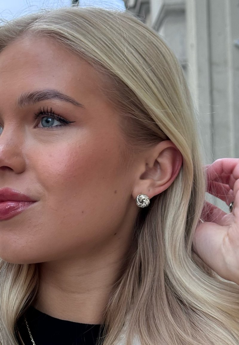 Silvriga knutstudsörhängen på en modell, med en texturerad design. Mjuk blondt hår faller förbi örat och framhäver accessoarerna.
