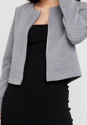 Femme portant une robe noire et une veste courte texturée gris clair avec un col rond, levant un bras vers sa tête.