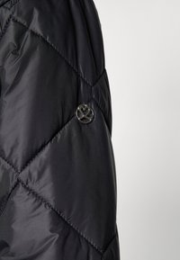 Crni puffer jakna s kvadratnim dijamantnim uzorkom, glatke teksture i kružnim logom s naglaskom s lijeve strane.