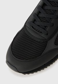 Sort atletiske sko med mesh- og gummimaterialer, rund tå, tekstureret overflade og flade snørebånd.