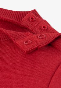 Szczegół czerwonego dzianego swetra z prążkowanym kołnierzem, dwoma okrągłymi czerwonymi guzikami na listwie oraz miękką, teksturowaną tkaniną.