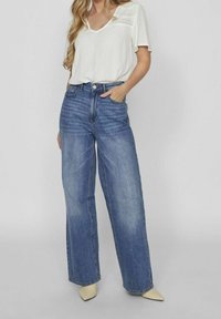 Femme portant un chemisier blanc à manches courtes avec des détails en dentelle, un jean bleu taille haute et large, et des chaussures beiges à bout pointu.