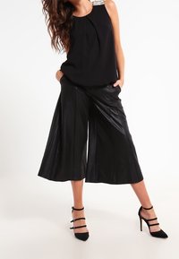 Top sans manches noir avec des plis et des bretelles ornées, associé à un pantalon culotte en faux cuir noir taille haute. Talons aiguilles noirs avec des lanières.