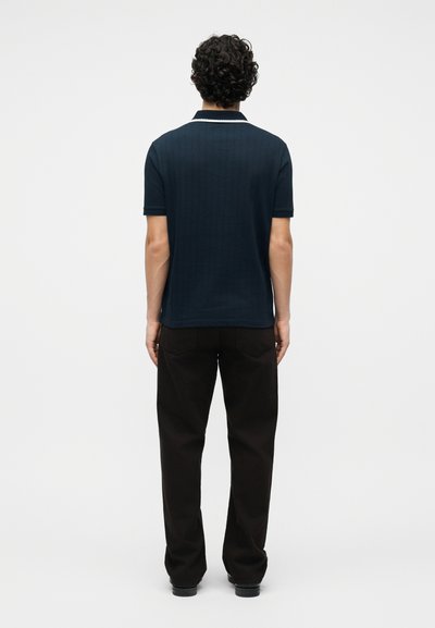 Άνδρας που φοράει μπλε σκούρο κοντομάνικο πόλο με λευκό περιθώριο στο γιακά και μαύρο παντελόνι, στέκεται με την πλάτη προς το μέρος μας μπροστά σε μονόχρωμο φόντο.