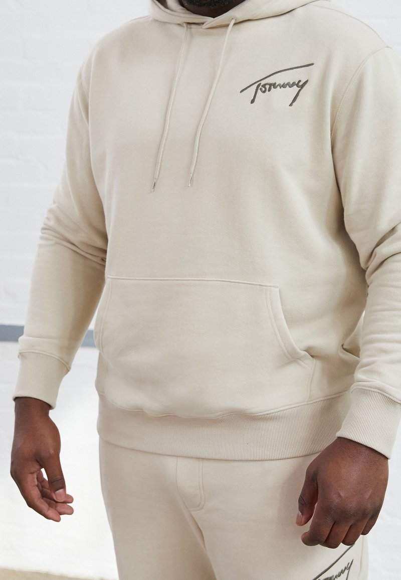 Homme portant un sweat à capuche beige et un pantalon assorti avec le logo « Tommy » sur la poitrine et la cuisse, debout avec une posture détendue.