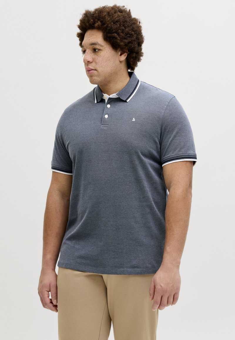 Mand med krøllet hår iført en grå polotrøje med hvide kanter og beige bukser, stående foran en ensartet lys baggrund.