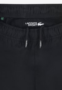 Black athletic shorts with an elastic waistband, featuring a mesh lining and two drawstrings; includes "LACOSTE SPORT" logo in white.