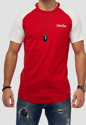 Red and white short-sleeve T-shirt with a crew neck, featuring a logo on the chest. Worn with distressed blue jeans and a dog tag necklace.