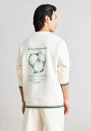 Mies, jolla on päällä off-white-värinen pusero vihreällä reunuksella, jossa vihreä jalkapallokuvio ja teksti "Scandinavian Football Community" selässä.