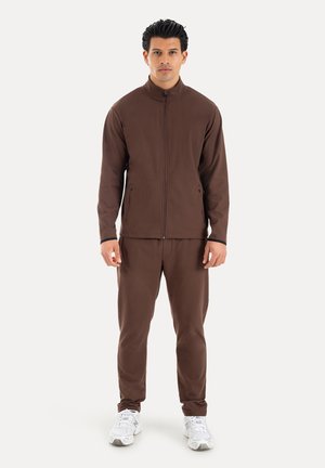 Homme debout face à nous, portant une veste marron zippée, un pantalon assorti et des baskets blanches sur un fond uni.