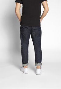 Vaqueros de mezclilla oscura con ajuste cónico, puños remangados y dos bolsillos traseros, combinados con zapatillas blancas y una camiseta negra.