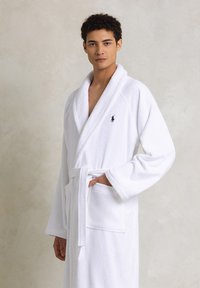 Accappatoio bianco in spugna con colletto a scialle, maniche lunghe e cintura. Include due tasche frontali e un piccolo logo ricamato.