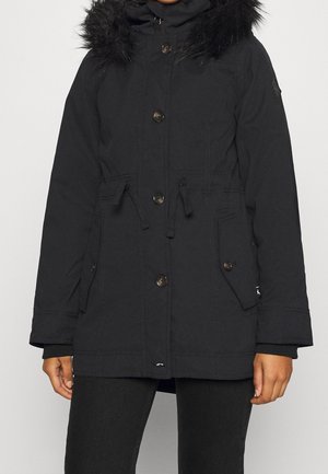 Parka d’hiver noire avec capuche bordée de fourrure, fermeture à boutons et zip, taille avec cordon de serrage et poches à rabat, portée par une personne debout.