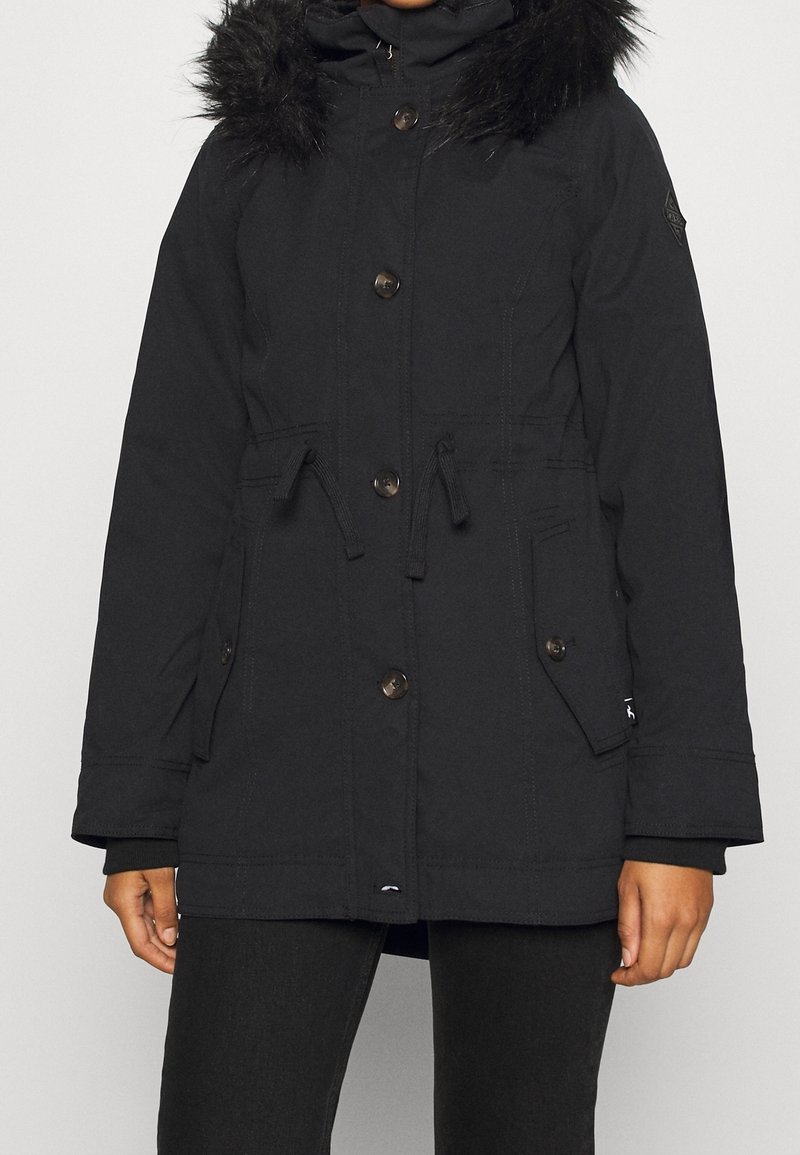 Parka d’hiver noire avec capuche bordée de fourrure, fermeture à boutons et zip, taille avec cordon de serrage et poches à rabat, portée par une personne debout.