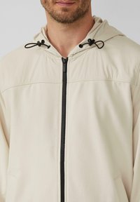 Giacca con cappuccio di un beige chiaro realizzata in tessuto liscio, con una cerniera nera, dettagli con cordini e un design semplice senza motivi.