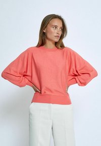 Korallrosa tröja med avslappnad passform, vida ärmar och ribbat avslut. Tillsammans med ljusfärgade, skräddarsydda byxor. Mjuk textur, enkel design.