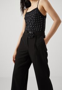 Kobieta w czarnej bezrękawnik z srebrnymi ćwiekami oraz czarnych spódnicach z wysokim stanem, z czarnym paskiem w róże, z rękami wsuniętymi w kieszenie.