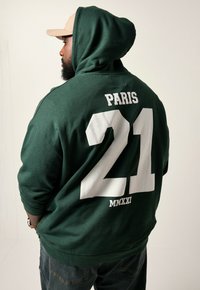 Grüne Kapuzenjacke mit weißem Drucktext "PARIS" und "21" sowie "MMXXI" darunter. Weiches Material, lockere Passform und eine große Fronttasche.