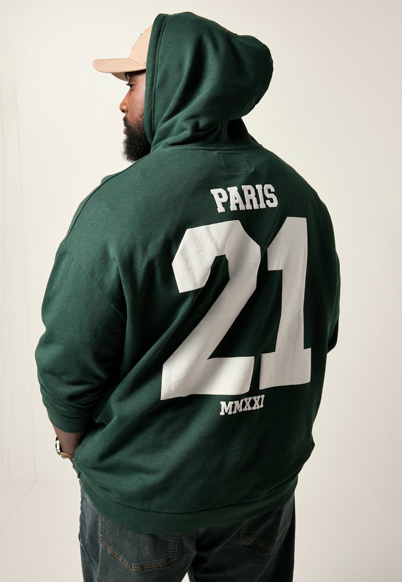 Grüne Kapuzenjacke mit weißem Drucktext "PARIS" und "21" sowie "MMXXI" darunter. Weiches Material, lockere Passform und eine große Fronttasche.