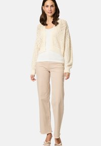 Crème gebreid vest met een cropped ontwerp, openwerkpatroon en aansluitende mouwen, gecombineerd met lichtbeige broeken en lichtgekleurde schoenen.