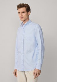 Camisa de botones de color azul claro en algodón, con un bolsillo en el pecho, botones blancos y mangas largas con detalles de puños abotonados.