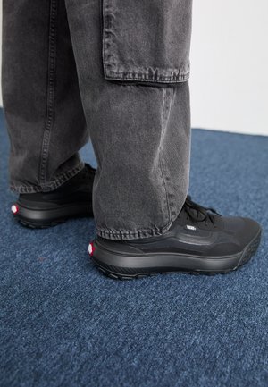 Personne portant des baskets Vans noires et un pantalon cargo gris, debout sur un sol moquetté bleu.