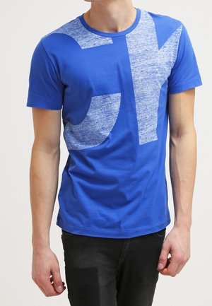 Mann trägt blaues kurzärmeliges T-Shirt mit großen hellblauen abstrakten Buchstaben und schwarze Hose, steht vor einem schlichten weißen Hintergrund.