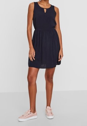 Femme portant une robe noire sans manches jusqu'aux genoux avec un décolleté en trou de serrure et des baskets décontractées roses, debout devant un fond uni.