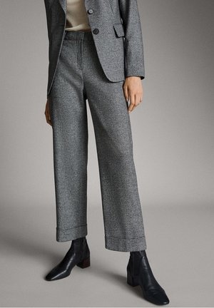 Woman wearing gray wide-leg trousers, matching gray blazer, beige top, and black ankle boots standing on a neutral background.