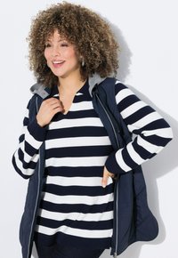 Marinblå pufferväst med grå huva, lager på lager över en vit och marinblå randig tröja. Modellen poserar med händerna i fickorna, vilket visar upp texturerna.