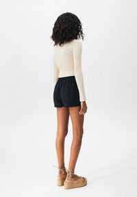 Kremowy prążkowany top z długim rękawem, czarne szorty z elastycznym pasem oraz jasnobrązowe sandały na koturnie z paskami, widziane od tyłu.