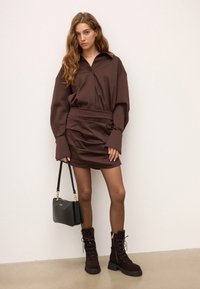 Mujer vistiendo un vestido mini de manga larga marrón, botas de cordones a juego, medias transparentes y sosteniendo un bolso negro, de pie contra una pared blanca.