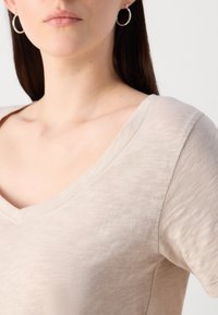 Top bej cu decolteu în V, realizat dintr-un material neted și texturat; mâneci scurte și detalii de cusătură curate. Include cercei mici, tip inel, ca accesoriu.