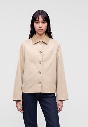 Donna con capelli lisci e scuri che indossa una giacca beige con chiusura con bottoni e maniche ampie, abbinata a jeans blu scuro.