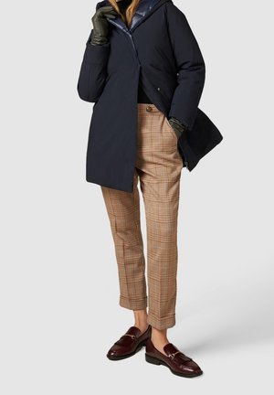 Frau trägt Navy-Mantel, karierte hochtaillierte Hose, grüne Lederhandschuhe und burgunderrote Loafers, steht vor einfarbigem Hintergrund.