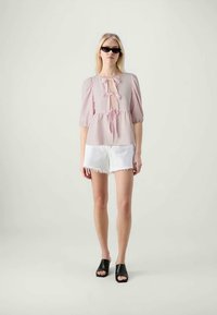 Helles rosa Bluse mit Puffärmeln und einer Schleife vorne, kombiniert mit weißen, zerrissenen Shorts und schwarzen, offenen Mules.