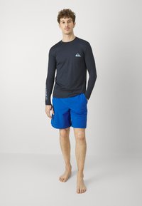 Čierny dlhý rukáv rash guard s malým logom na hrudi, v kombinácii s živými modrými plavkami. Mäkko tkanina a voľný strih, minimálne akcenty.