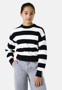 Pull en rayures noir et blanc avec texture côtelée, encolure ronde et manches longues, associé à un pantalon gris clair.