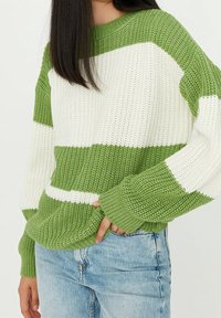 Donna che indossa un maglione a righe verdi e bianche con jeans azzurri, con la mano destra in tasca, in piedi contro uno sfondo neutro.