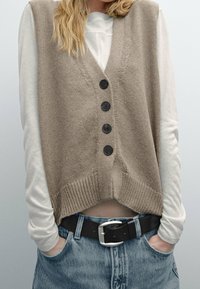 Beige neuleliivi, jossa on V-pääntie, viisi mustaa nappia ja ribattu helma, kerrostettuna vaalean pitkähihaisen paidan kanssa ja yhdistettynä farkkushortsien kanssa.