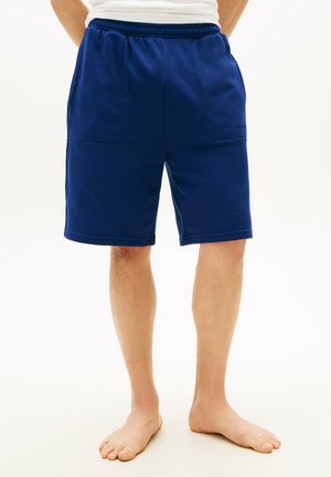 Barfuß stehende Person, die dunkelblaue, knielange Shorts mit Vorderentaschen und ein weißes Oberteil trägt, vor einem einfarbigen Hintergrund.