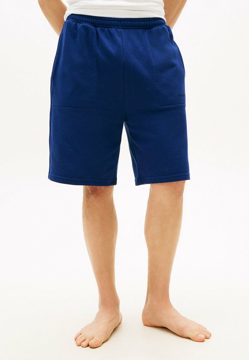 Personne pieds nus portant un short bleu foncé jusqu'aux genoux avec des poches avant et un haut blanc, debout devant un fond uni.