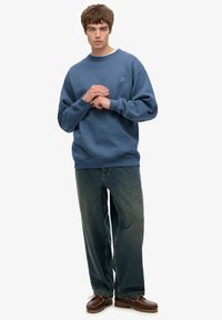Blauwe sweatshirt met ribbelkraag, gecombineerd met losse, donkere spijkerbroek en bruine schoenen. Eenvoudig ontwerp, geen zichtbare patronen of afbeeldingen.