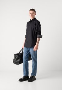 Svart skjorte med knapper og mønstrede mansjetter, blå denim jeans og svarte skinnsko. Holder en svart skinn duffelbag.