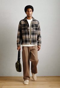 Plaid flannel shirt in brown and beige, white t-shirt, brown wide-leg pants, and light sneakers. Carrying a small, olive green bag.